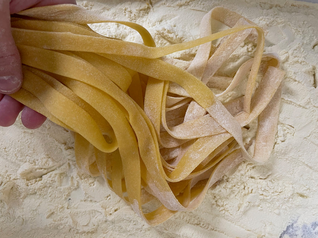 Tagliatelle pasta arranged in a nest shape on a wooden surface with flour dusted around it. The pasta strands are long and flat, with a slightly yellow hue, and have a slightly curled edge. Light shadows are visible on the surface, indicating the pasta's texture